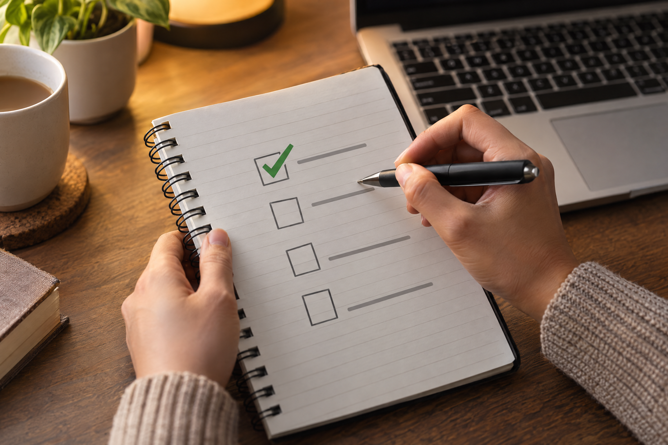 Hands reviewing a checklist in a notebook beside a laptop, with one item ticked and one waiting for approval